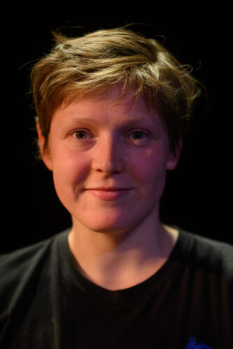 A white woman with short light brown hair. She is wearing a black t-shirt and is smiling at us.