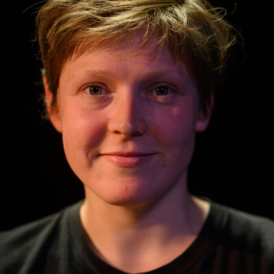 A white woman with short light brown hair. She is wearing a black t-shirt and is smiling at us.