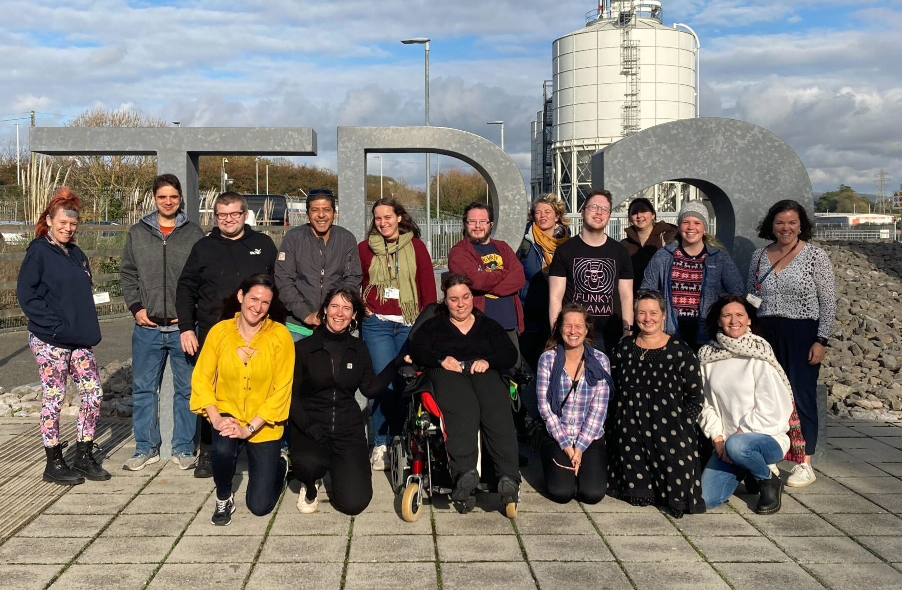 Outdoors setting, half blue sky, half clouds. A diverse group of lovely people stand, crouch and sit in wheelchairs together for a group photo. They're all in front of a large 3D sign that reads "TR2".