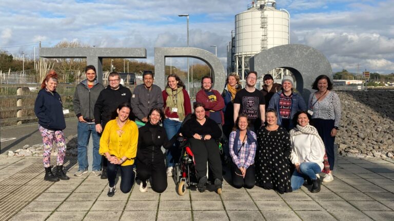 Outdoors setting, half blue sky, half clouds. A diverse group of lovely people stand, crouch and sit in wheelchairs together for a group photo. They're all in front of a large 3D sign that reads "TR2".