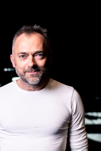 Headshot of a white man with a greying stubble beard, short hair and bright blue eyes. He wears a white tight top and smiles softly at the camera.