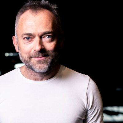 Headshot of a white man with a greying stubble beard, short hair and bright blue eyes. He wears a white tight top and smiles softly at the camera.