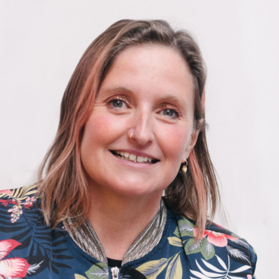 A white women with shoulder-length brown and faded-pink hair smiles at the camera. She wears a nose ring and a tropical-print bomber jacket.