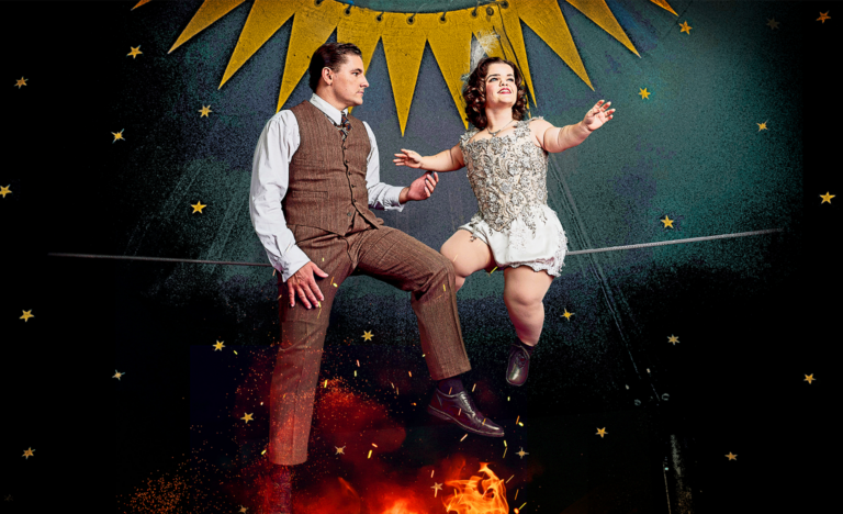 The main image is two performers sat on a raised tight wire in a dark circus tent. The circus tent is decorated with gold stars and bunting. To the right is the main character, Krista who is a young white woman of short stature with short brown hair looking away into the distance. To the left is Gerhard, a young white man with short brown hair, who gazes intently at the other performer, Krista, as he reaches his hand to touch her. Both characters’ costumes are in the style of the 1930s. Hers is a shiny silver beaded corset with white silk shorts, tights and black shoes. He looks elegant in brown trousers and a brown waistcoat over a long-sleeved white shirt and black shoes. Underneath the two performers burns a small fierce fire with red flames, reaching the soles of their feet and signifying the danger beneath this love story" Photo credit: Paul Blakemore Design by Studio Stiles