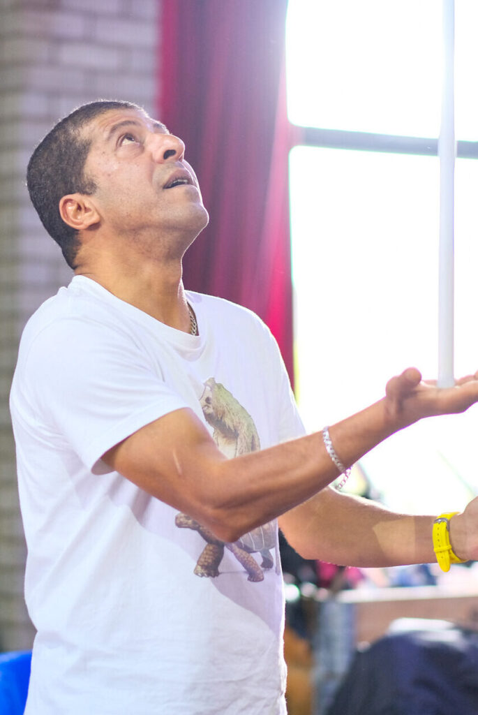 A Brown man with short cropped hair and a white T-shirt balances a white stick on the palm of his hand. He stares at it with a look of concentration on his face.