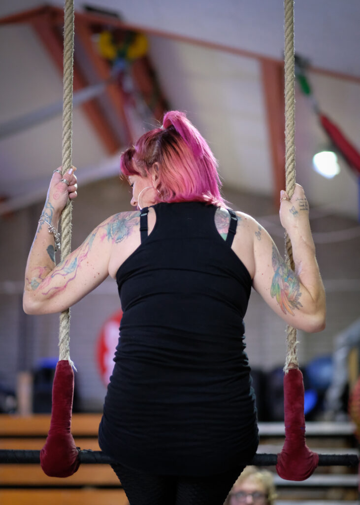 A white woman with her back to us. She wears a black tank op and has bright pink shoulder-length hair, and blue and red tattoos on her arms. She is sitting on a trapeze and looking to her bottom left. Her hands grip the trapeze ropes.