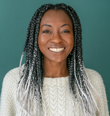 Beryl is a young Black woman with a bright smile. She has long braids, their top half is black and their lower half is white. She wears a cream jumper and is posing in front of a teal background.