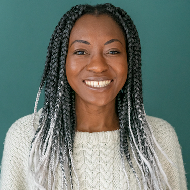 Beryl is a young Black woman with a bright smile. She has long braids, their top half is black and their lower half is white. She wears a cream jumper and is posing in front of a teal background.