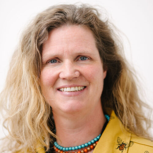 Headshot of Paula, a white woman in her forties with long wavy blonde hair that falls just below her shoulders. She wears a yellow denim top with bees on it and turquoise and orange beads around her neck. She's smiling warmly at the camera.