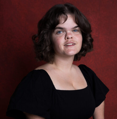 Headshot of a white woman with shoulder-length wavy brown hair and blue eyes. She wears a short sleeve black dress. She is softly smiling at the camera.