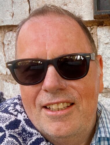 Middle aged white man wearing sunglasses and checked shirt. He is smiling at the camera.