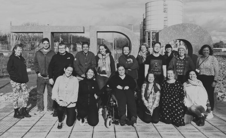 Black and white image. Outdoors setting. A diverse group of lovely people stand, crouch and sit in wheelchairs together for a group photo. They're all in front of a large 3D sign that reads "TR2".