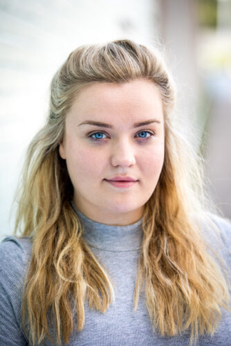 An outdoors headshot of a white woman with long blonde hair worn half up. She is wearing a grey turtleneck.