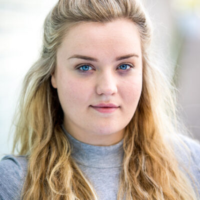 An outdoors headshot of a white woman with long blonde hair worn half up. She is wearing a grey turtleneck.