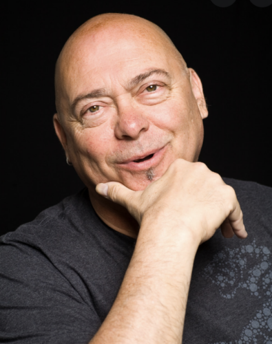 White man with a goatee and clean shaven head. He is wearing a grey t-shirt and has his hand on his chin.