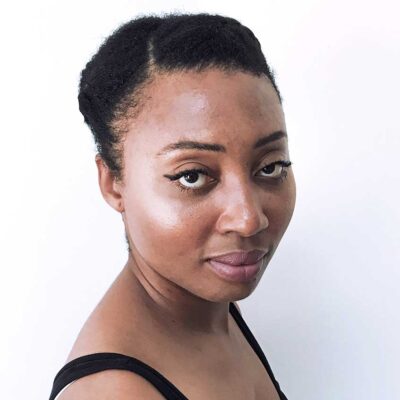 Headshot of Grace, a Black woman with dark brown eyes and hair pinned back tightly on her head. She wears a black tank top and black eyeliner above her eyes.