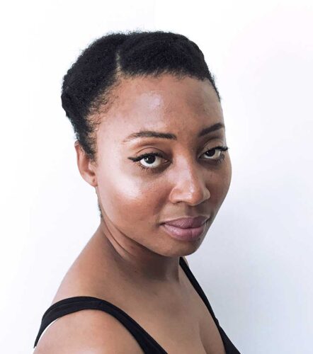 Headshot of Grace, a Black woman with dark brown eyes and hair pinned back tightly on her head. She wears a black tank top and black eyeliner above her eyes.