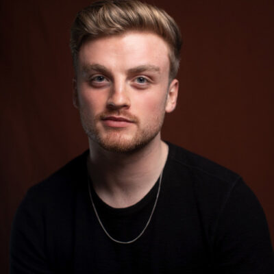 An indoor headshot of a white man with short strawberry blonde hair and facial hair. He is wearing a black t-shirt and a thin necklace.