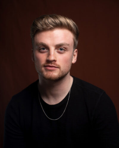 An indoor headshot of a white man with short strawberry blonde hair and facial hair. He is wearing a black t-shirt and a thin necklace.