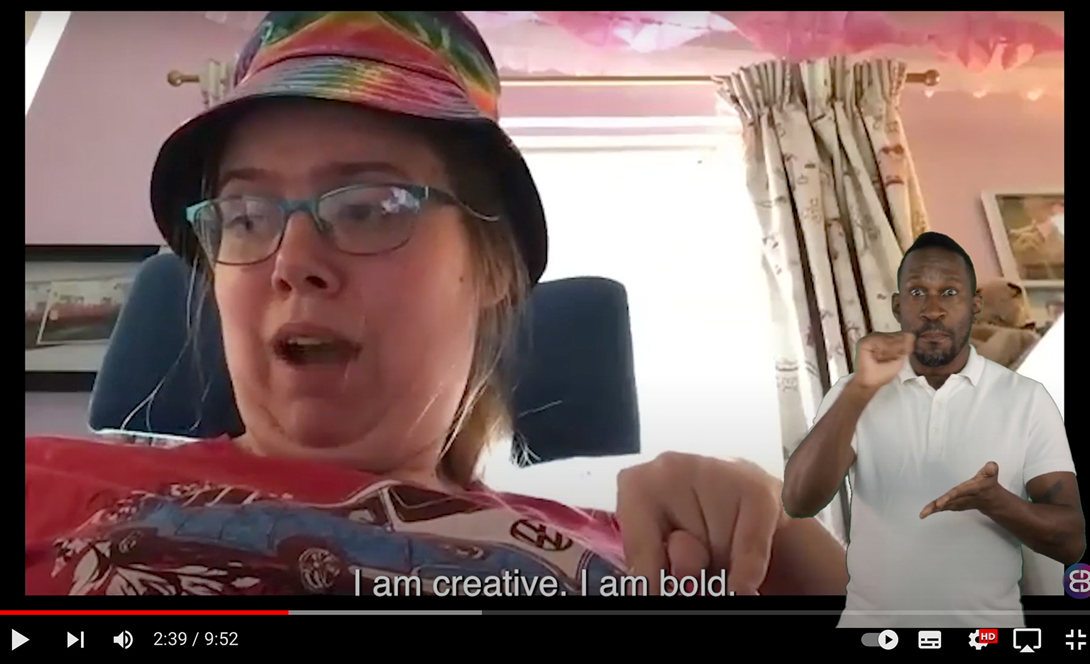 Screenshot of a film playing on a screen. A black and red playbar at the bottom shows we're about one third into the video. The video shows a white person sitting in a wheelchair. She wears a multicoloured tie and dye bucket hat and a colourful patterned top speaking to the camera. A caption below them reads: "I am creative. I am bold.".