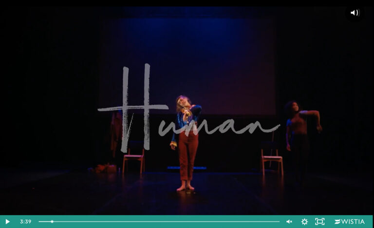 Screenshot of a film playing on a screen. A teal-coloured play bar at the bottom shows we're almost at the end of a long video. The video shows a dimly-lit stage with two performers standing. One is in the centre in the foreground. She stand with one arm raised up to grab a trapeze. The other one is in the background to the right. She is standing with her arms extended out to the side. A title superimposed on the screen, in handwritten white letters reads: "Human".