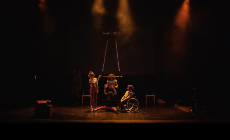 A dimly lit stage, soft orange light and smoke. Four performers together in the centre. One performer is sat on the floor with their legs splayed out. They're holding onto a trapeze. The others around them are comforting them and helping them let go of the trapeze bar.