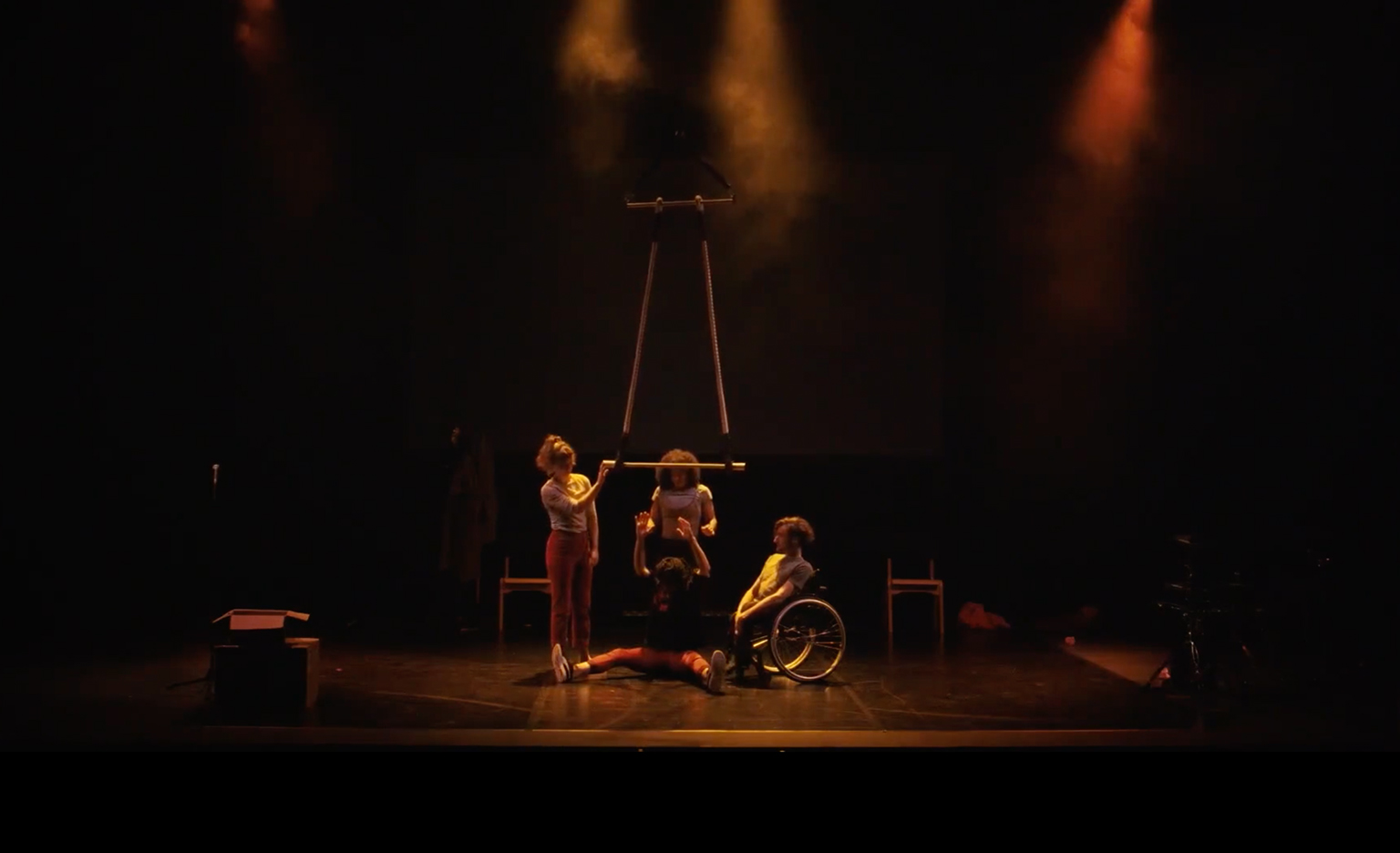 A dimly lit stage, soft orange light and smoke. Four performers together in the centre. One performer is sat on the floor with their legs splayed out. They're holding onto a trapeze. The others around them are comforting them and helping them let go of the trapeze bar.