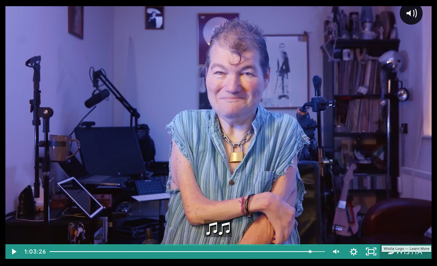 Screenshot of a film playing on a screen. A teal-coloured play bar at the bottom shows we're almost at the end of a long video. The video shows a white man wearing a light blue striped short-leeved shirt, a thick padlock on a chain around his neck and hair in a signature Kiss curl. Behind him, music kit and instruments and a living room setting.