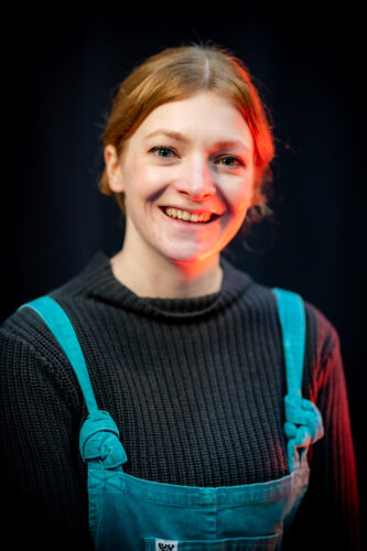 Young white woman with ginger hair tied back. She is wearing a black longsleeve t-shirt with turquoise dungarees. She is smiling at the camera.