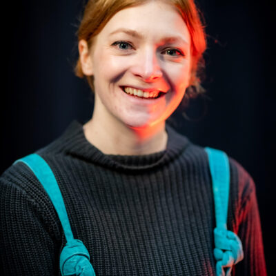 Young white woman with ginger hair tied back. She is wearing a black longsleeve t-shirt with turquoise dungarees. She is smiling at the camera.