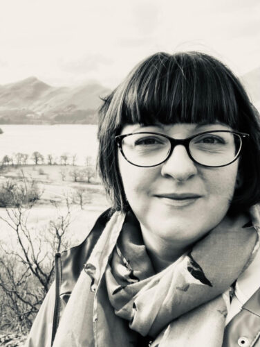 Black and white photo of a white woman wearing glasses. She has brown hair with a fringe and bob. She is wearing a scarf. Mountains and a lake can be seen in the background.