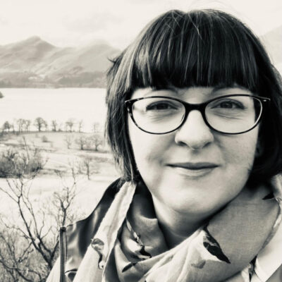 Black and white photo of a white woman wearing glasses. She has brown hair with a fringe and bob. She is wearing a scarf. Mountains and a lake can be seen in the background.