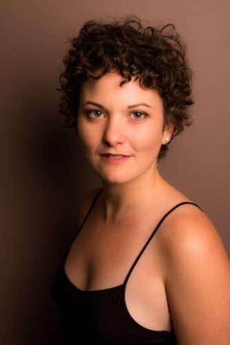 Sepia toned headshot of a white woman with short wavy brown hair. She is wearing a black tank top and is looking at the camera with a soft smile.