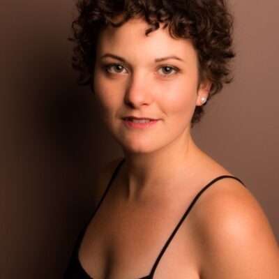 Sepia toned headshot of a white woman with short wavy brown hair. She is wearing a black tank top and is looking at the camera with a soft smile.