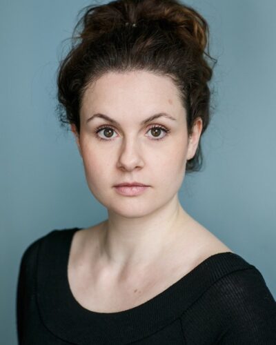 Headshot of a young white woman wearing a black long-sleeve t-shirt. She has curly brown hair which is worn in an up-do.