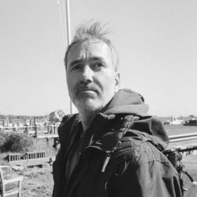 Headshot of Glenn, a white man in his forties with stubble beard and greying hair. He's wearing rain jacket and a backpack. He's standing outside with houses and grass and benches behind him.