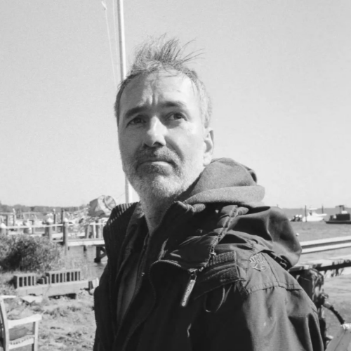 Headshot of Glenn, a white man in his forties with stubble beard and greying hair. He's wearing rain jacket and a backpack. He's standing outside with houses and grass and benches behind him.