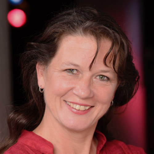 Headshot of Sula, a white woman in her forties with long wavy brown hair tied back, blue eyes and lightly pink-painted lips. She wears a red blouse and smiles at the camera.