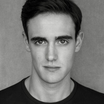 Black and white headshot of a white man with black hair looking charismatically at the camera. He is wearing a black tshirt.