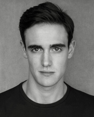 Black and white headshot of a white man with black hair looking charismatically at the camera. He is wearing a black tshirt.