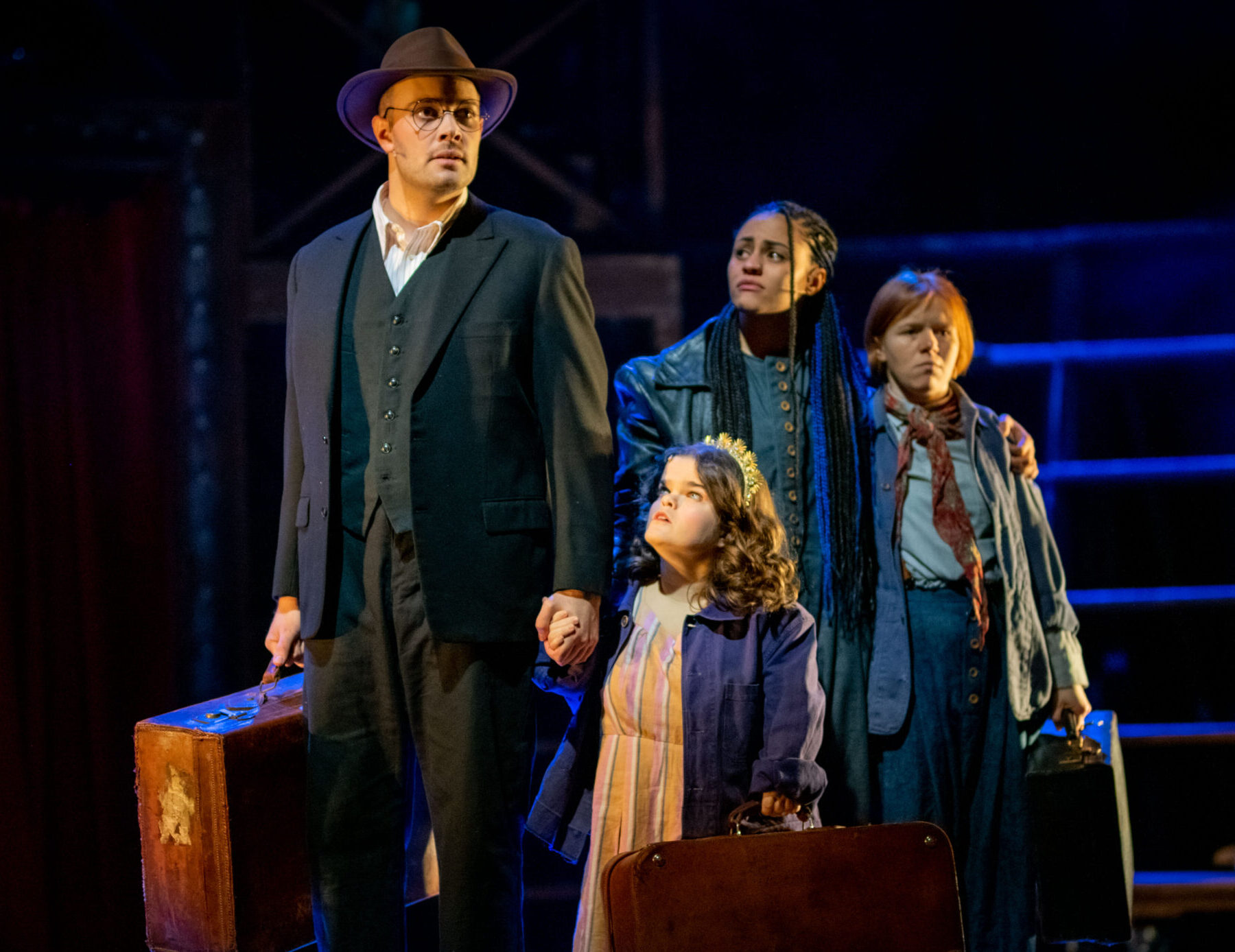 Dimly lit stage is illuminated by blue lights. Four performers onstage holding briefcases. they all look worried as they look out into the audience.