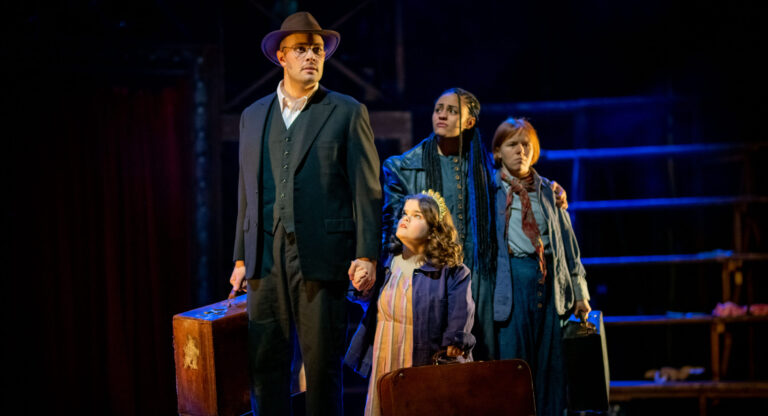 Dimly lit stage is illuminated by blue lights. Four performers onstage holding briefcases. they all look worried as they look out into the audience.