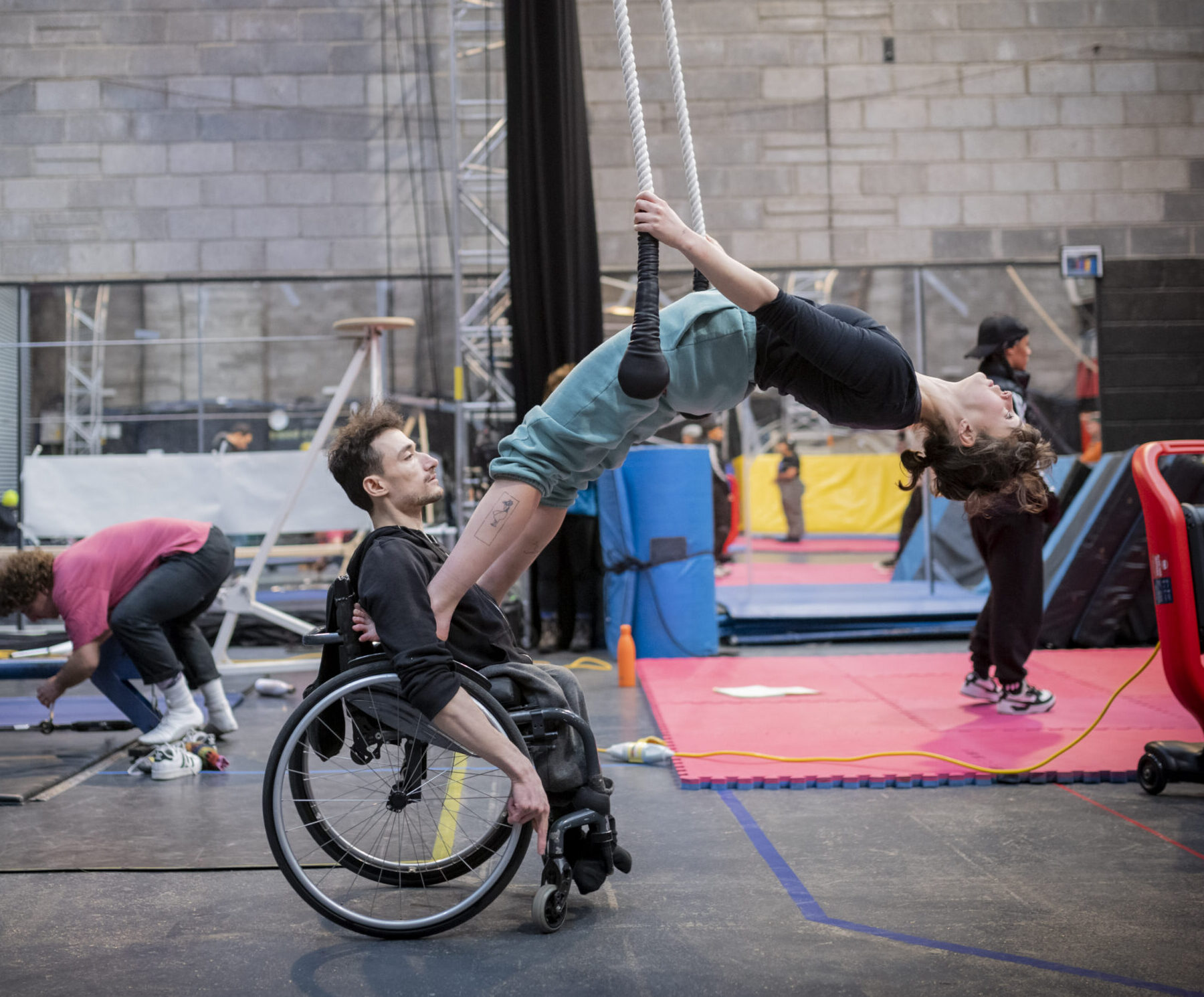 Indoor rehearsal room with high ceilings. Mats and rigging can be seen in the background. Two performers rehearsing a routine. The first performer is a white man with brown hair. He is a wheelchair user. The other performer is a white woman with brown hair. She has her feet on the handles of the wheelchair and is leaning back onto an aerial trapeze.