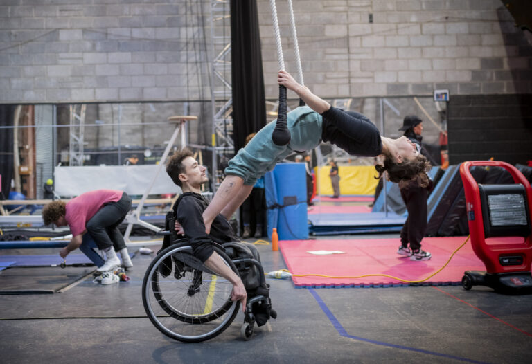 Indoor rehearsal room with high ceilings. Mats and rigging can be seen in the background. Two performers rehearsing a routine. The first performer is a white man with brown hair. He is a wheelchair user. The other performer is a white woman with brown hair. She has her feet on the handles of the wheelchair and is leaning back onto an aerial trapeze.