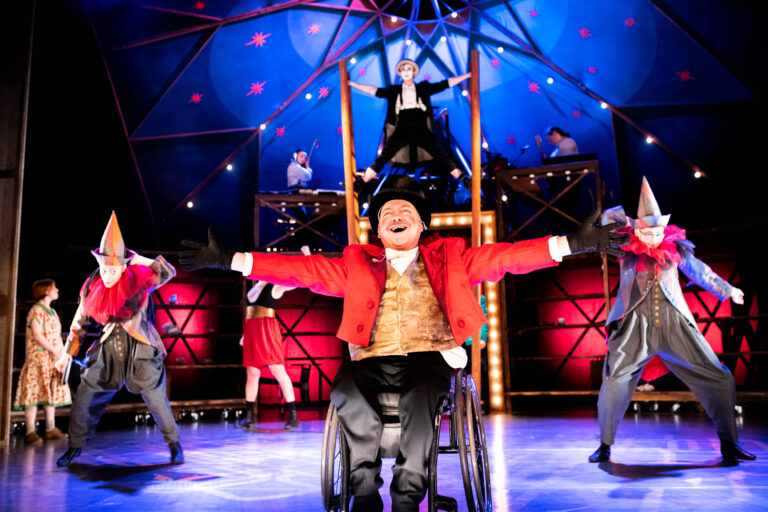 A colourful blue and red stage with a circus top as the scenery. There are multiple performers onstage wearing colourful and vibrant costumes, all circus themed. To the forefront is a white man in a wheelchair wearing a ringmaster costume with a top hat. He has his arms outstretched and has a big smile on his face.