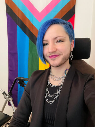 Indoor office space. A person is sat in a office chair and is smiling at the camera. They have blue hair and is wearing a brown blazer over a black tshirt.