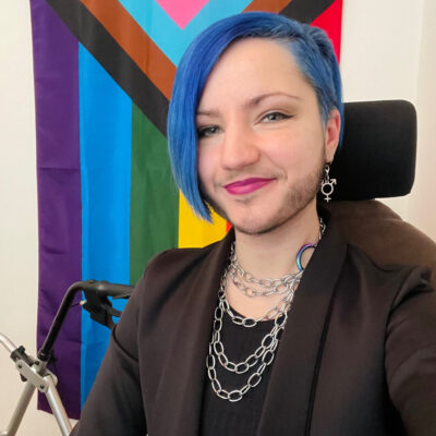 Indoor office space. A person is sat in a office chair and is smiling at the camera. They have blue hair and is wearing a brown blazer over a black tshirt.