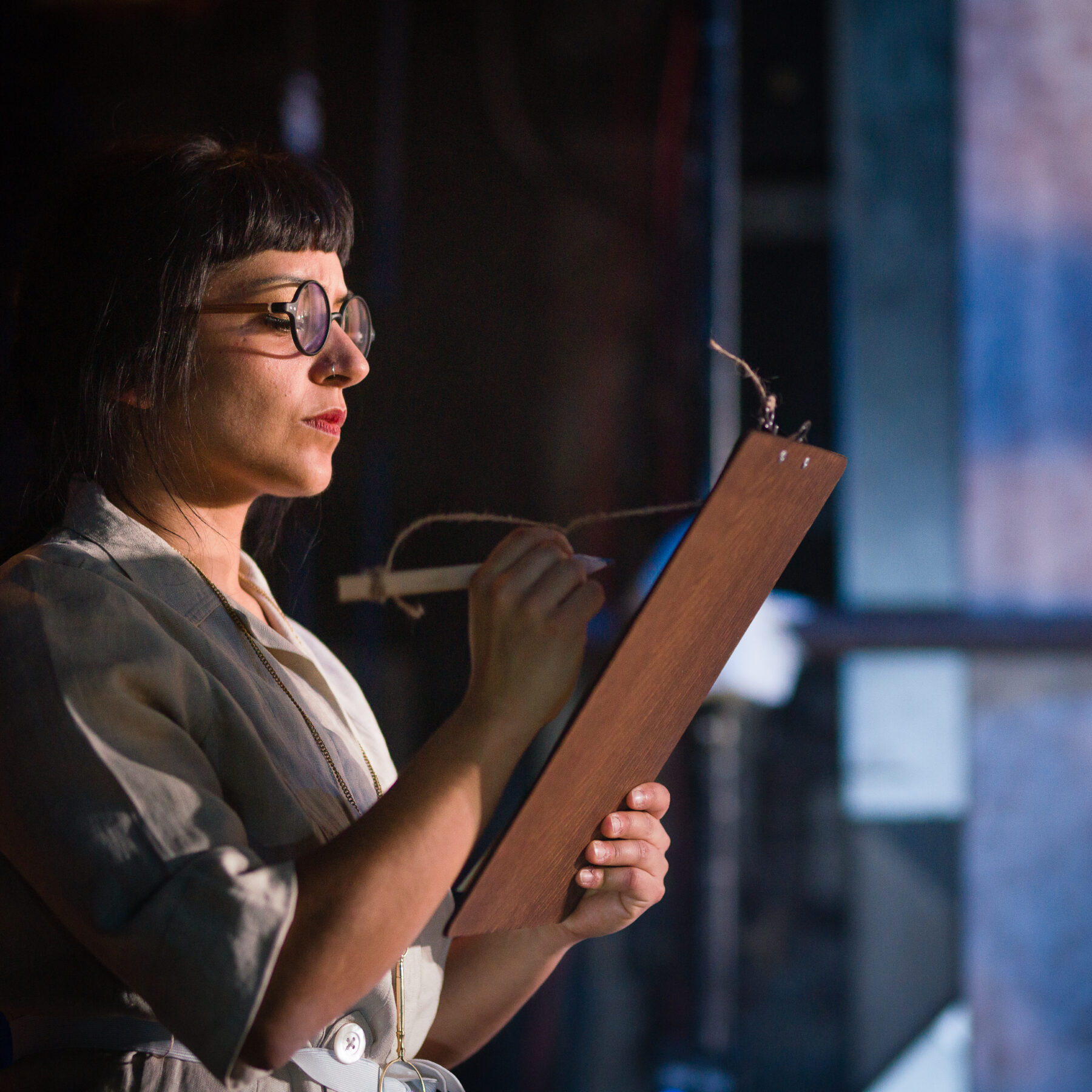 Dimly lit stage. The backstage area is slightly in view. A performer is holding a clipboard and writing on it very sternly. The performer is a woman of colour wearing glasses. She has black hair that is tied back with a short fringe.