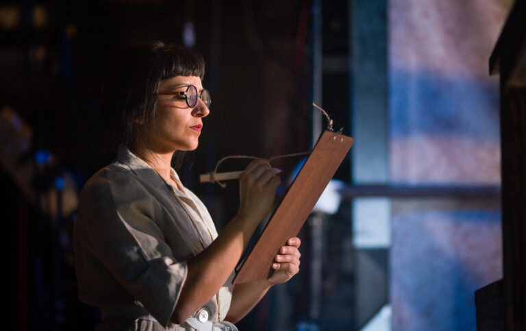 Dimly lit stage. The backstage area is slightly in view. A performer is holding a clipboard and writing on it very sternly. The performer is a woman of colour wearing glasses. She has black hair that is tied back with a short fringe.