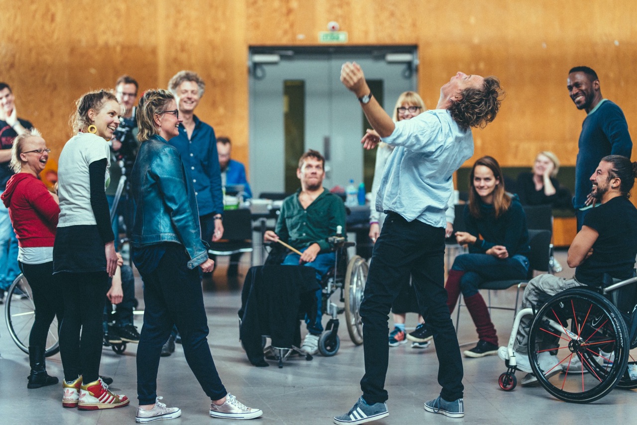 The composer, leans back with his head dramatically as the other members of the group rehearsal, who are gathered in a circle, some standing and some seated, look at him with smiles and laughing expressions.